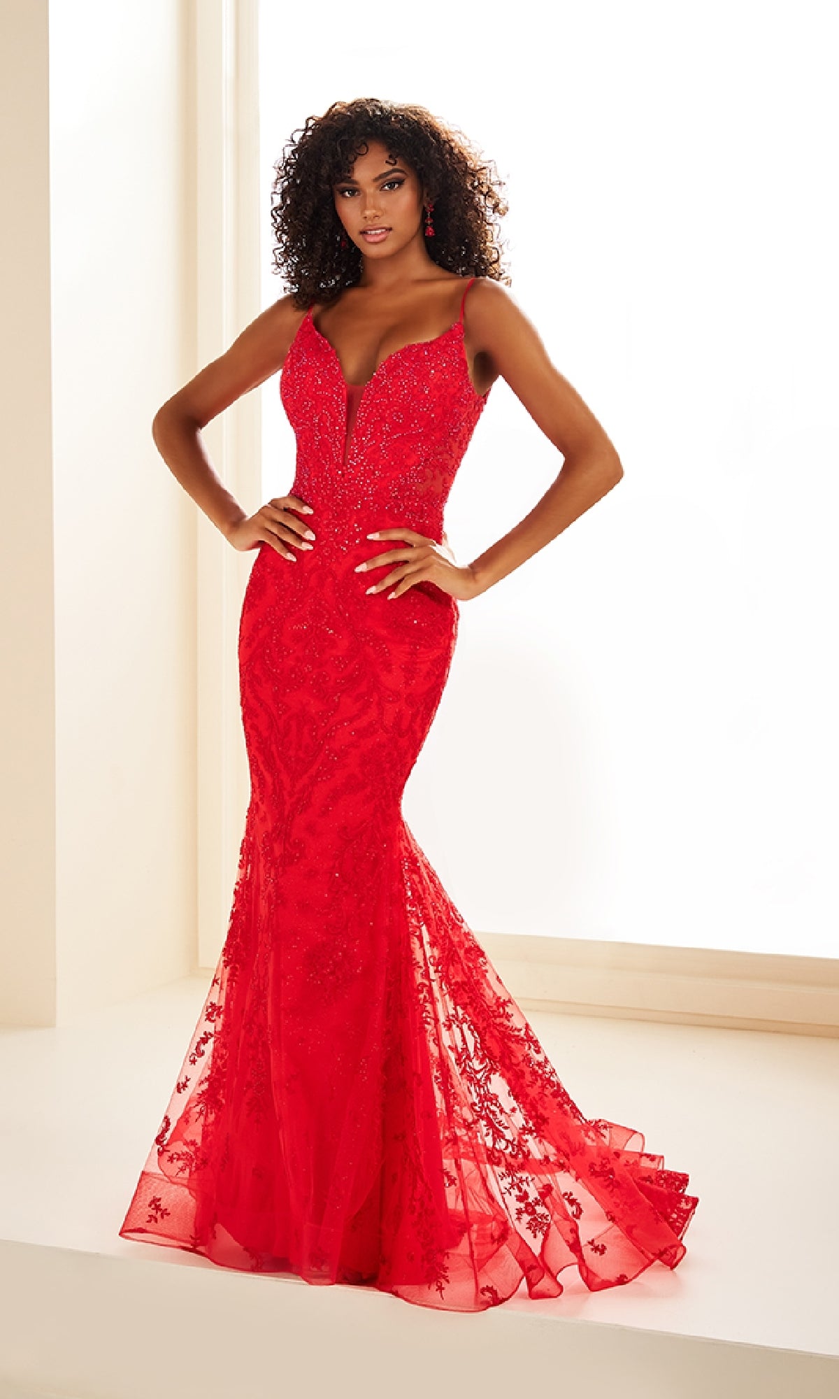 A woman poses hands on hips in the Ellie Wilde EW36096, a fitted floor-length mermaid prom dress featuring lace embroidery, a deep V-neckline, and thin straps, set against a white background.